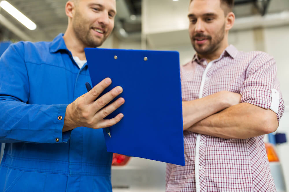 auto service, repair, maintenance and people concept - mechanic with clipboard talking to man or owner at car shop